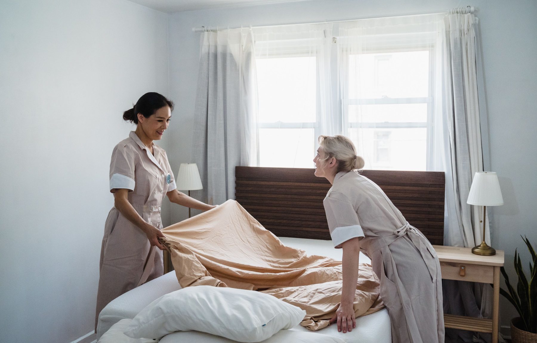 Zwei Pflegekraefte beziehen gemeinsam laechelnd ein Bett in einem hellen Zimmer
